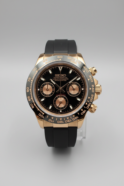 A Seiko Daytona watch with a rose gold-toned case, black dial, and black rubber strap, photographed from above on a dark gray background.