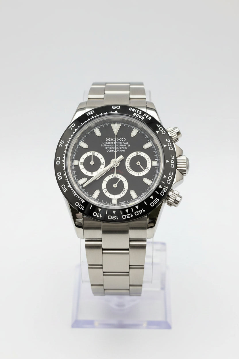 A Seiko Daytona watch with a dark gray dial, silver-tone stainless steel case and bracelet, and a black tachymeter bezel, displayed on a clear stand against a black background.