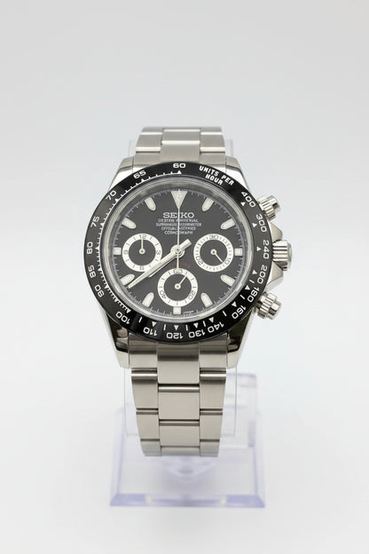 A Seiko Daytona watch with a dark gray dial, silver-tone stainless steel case and bracelet, and a black tachymeter bezel, displayed on a clear stand against a black background.