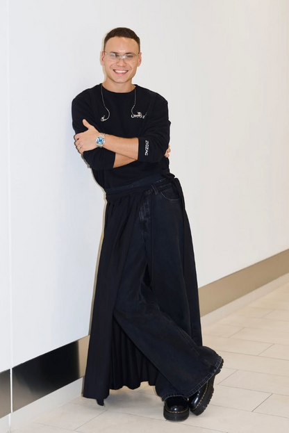A man wearing a black long-sleeve shirt and wide-leg black trousers, leaning against a white wall in a bright indoor space.