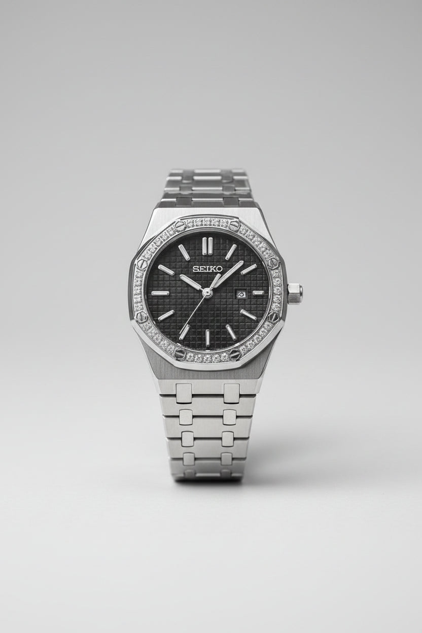 A Seiko stainless steel watch with a diamond-encrusted bezel, black textured dial, and matching metal link bracelet, shown close-up on a light fabric surface.