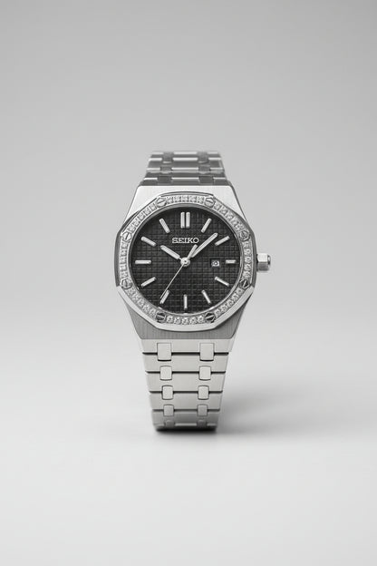 A Seiko stainless steel watch with a diamond-encrusted bezel, black textured dial, and matching metal link bracelet, shown close-up on a light fabric surface.