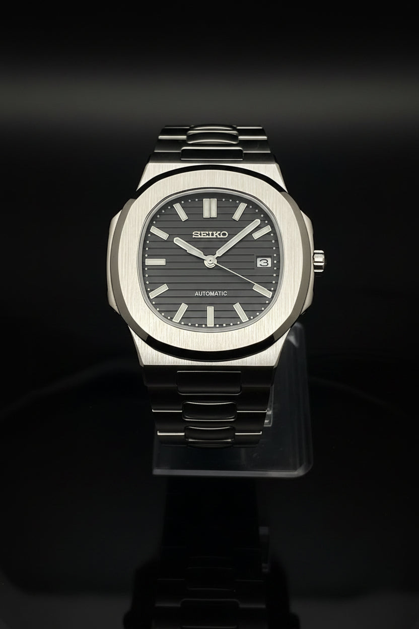 A Seiko automatic wristwatch with a brushed silver-tone metal case and integrated metal bracelet, featuring a dark gray dial with silver-tone hour markers and hands, displayed on a clear stand against a dark background.
