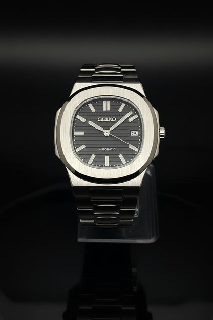 A Seiko automatic wristwatch with a brushed silver-tone metal case and integrated metal bracelet, featuring a dark gray dial with silver-tone hour markers and hands, displayed on a clear stand against a dark background.