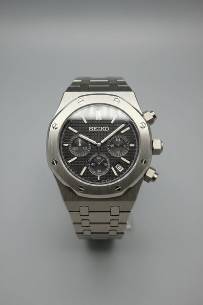 A Seiko wristwatch with a dark textured dial, a silver-toned metal case, and a matching metal link bracelet, photographed against a dark black background.