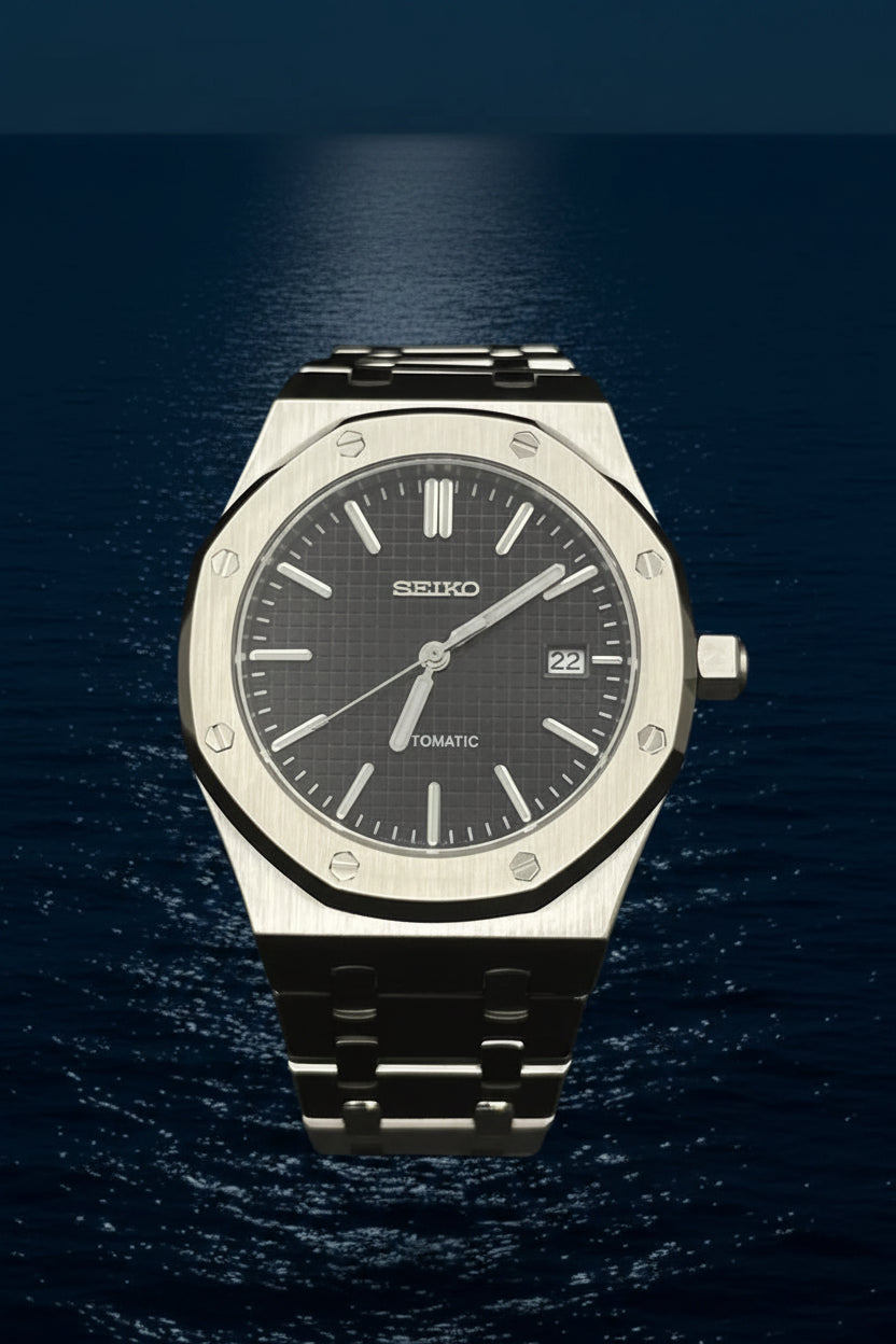 A Seiko automatic wristwatch with a brushed stainless steel case, octagonal bezel, and a dark gray dial, displayed on a clear stand against a black background.