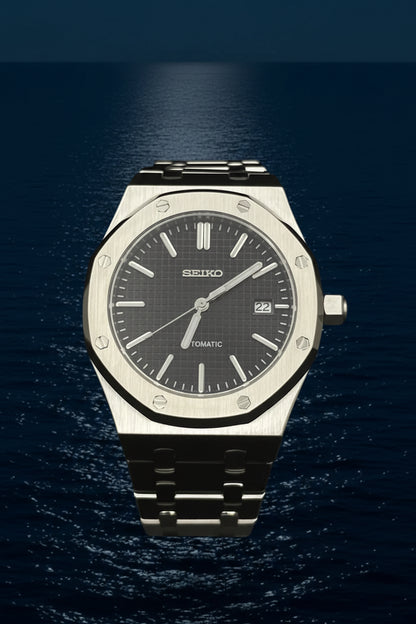 A Seiko automatic wristwatch with a brushed stainless steel case, octagonal bezel, and a dark gray dial, displayed on a clear stand against a black background.