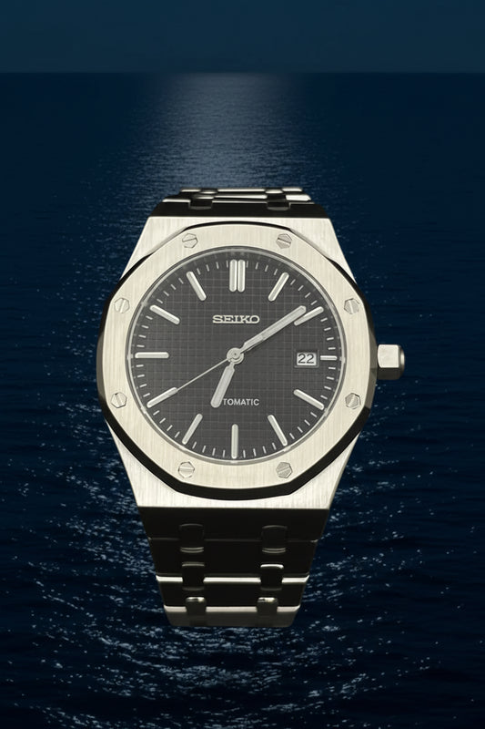 A Seiko automatic wristwatch with a brushed stainless steel case, octagonal bezel, and a dark gray dial, displayed on a clear stand against a black background.