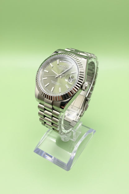 A Seiko stainless steel watch with a fluted bezel and a green dial, displayed on a clear stand against a dark gray background.