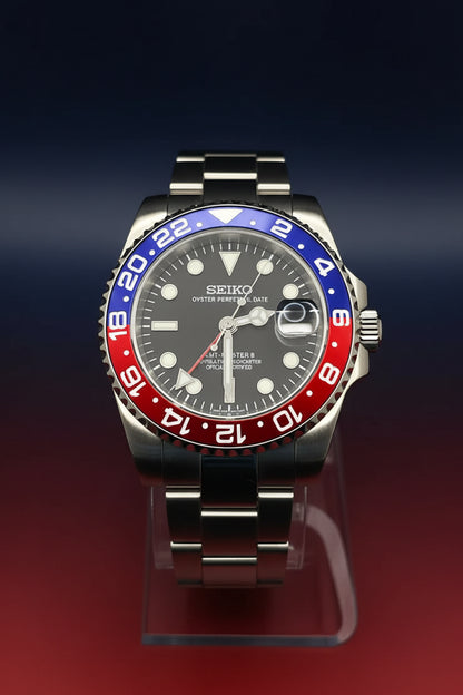 A Seiko Steel Ocean Perpetual watch with a stainless steel case, dark gray dial, and a distinctive red and blue GMT bezel, displayed on a clear stand against a black background.