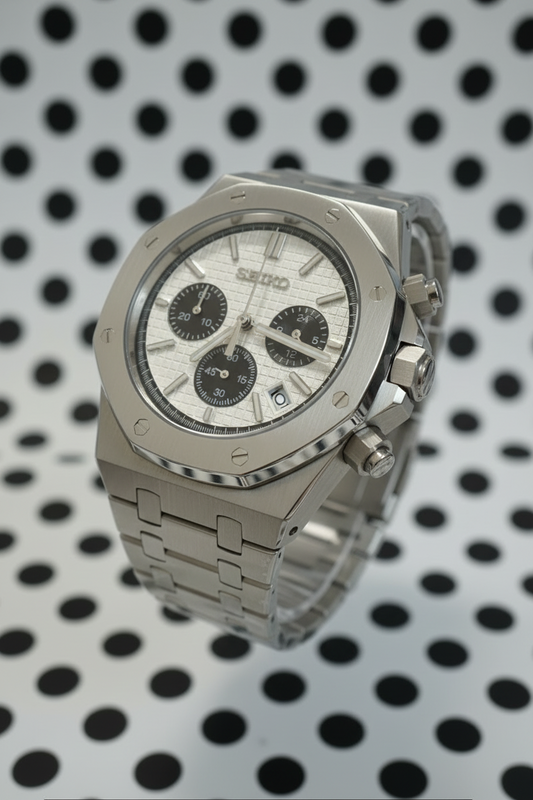 A stainless steel chronograph watch with an octagonal bezel, silver textured dial, and matching metal link bracelet, resting on a gray textured surface.