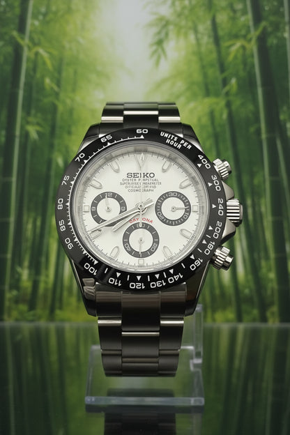 A Seiko Daytona watch with a silver stainless steel case, white dial, and black tachymeter bezel, displayed on a clear stand against a black background.